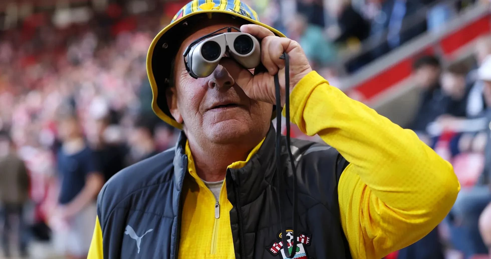 A fan wearing binoculars and a hat at the stadium